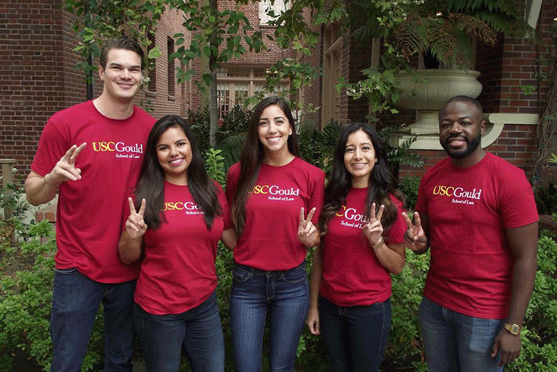 Five students wearing cardinal colored t-shirts printed with the USC Gould logo smile and make the Fight On gesture.