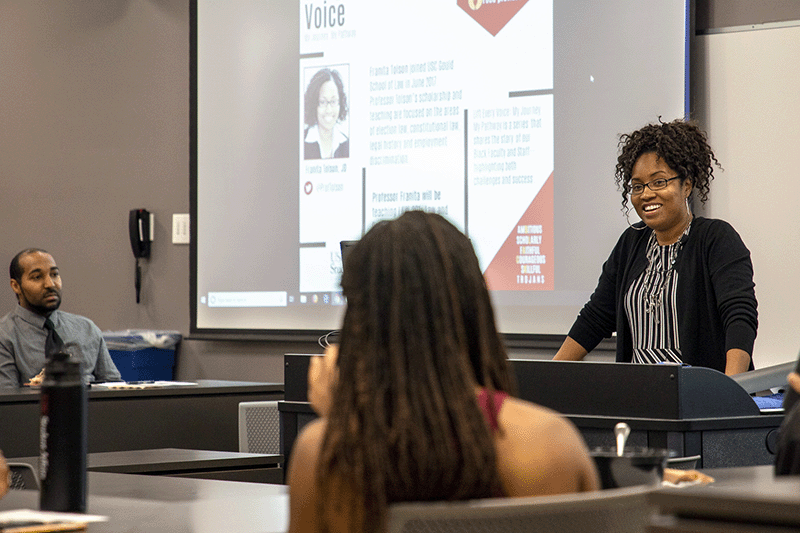 Professor Franita Tolson smiles from a podium, in front of a projected screen, facing undergrads participating in her Lift Every Voice lecture.