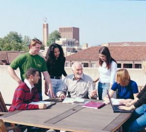 Professor Michael Brennan sits outside while collaborating with six PCJP students, smiling.