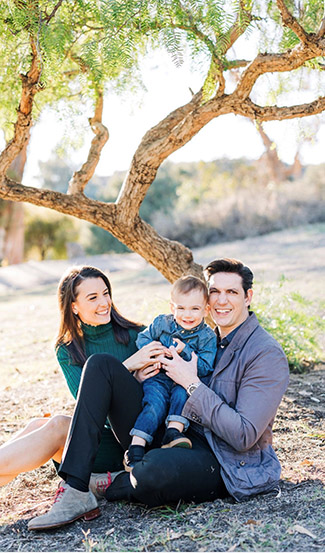 LLM alumnus, Moises Amsel, sits under a tree smiling with his wife and child.