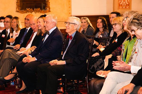 Professor Alex Capron sits with an audience in the front row, right seat, at an event celebrating his retirement.