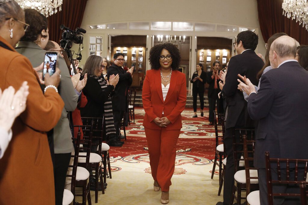 A woman in a red suit walks down a center aisle as the audience stands and claps for her.